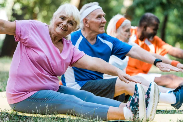 Focus selettivo di uomini e donne pensionati multiculturali in abbigliamento sportivo seduti su tappeti fitness — Foto stock