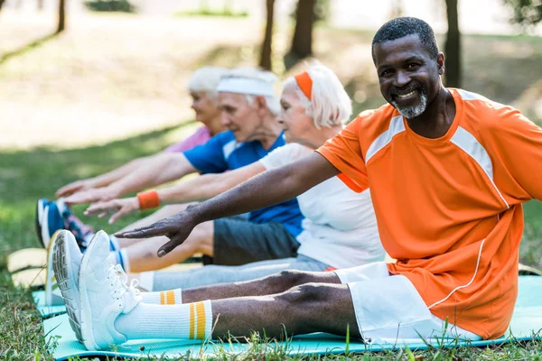 Enfoque selectivo de hombre afroamericano feliz cerca de jubilados pensionistas en ropa deportiva ejerciendo en colchonetas de fitness - foto de stock