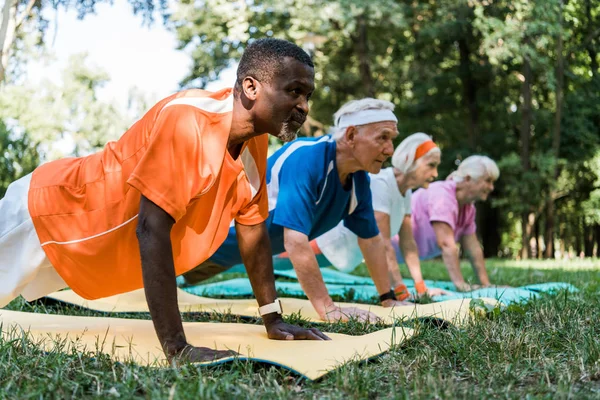 Enfoque selectivo del hombre afroamericano haciendo ejercicio de tablón con pensionistas jubilados en colchonetas de fitness - foto de stock