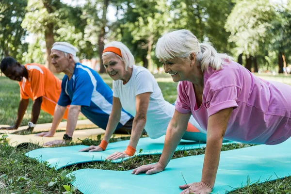 Focalizzazione selettiva della donna anziana felice che fa esercizio di asse con pensionati multiculturali su stuoie fitness — Foto stock