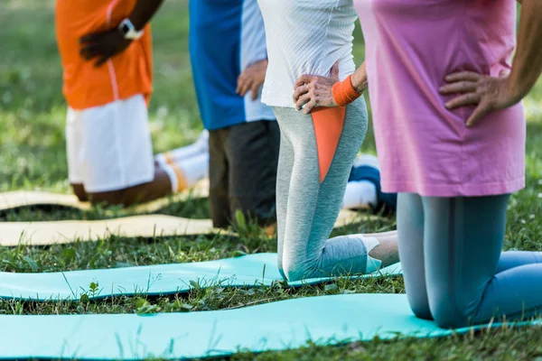 Vista cortada de pensionistas multiculturais com a mão em quadris exercitando-se em tapetes fitness — Fotografia de Stock