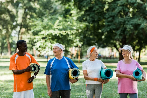 Heureux retraités âgés et multiculturels tenant des tapis de fitness tout en se tenant dans le parc — Photo de stock