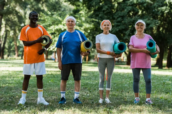 Aposentados felizes e multiculturais pensionistas segurando tapetes de fitness enquanto em pé na grama verde no parque — Fotografia de Stock