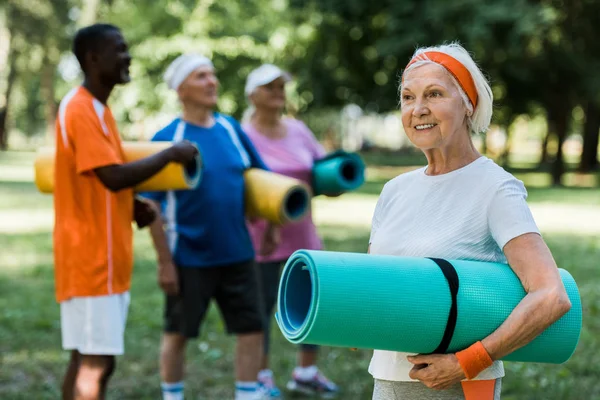 Selective focus of happy retired woman holding fitness mat near multicultural pensioners — Stock Photo