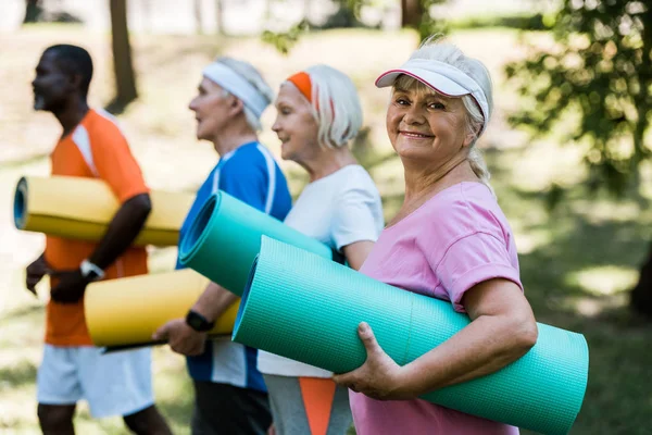 Foco seletivo da mulher aposentada feliz em cap segurando tapete fitness perto de pensionistas multiculturais — Fotografia de Stock