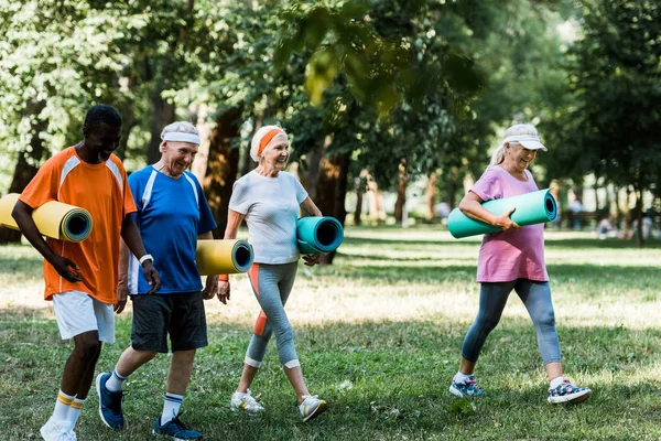 Enfoque selectivo de felices pensionistas multiculturales y senior caminando con colchonetas de fitness en el parque - foto de stock