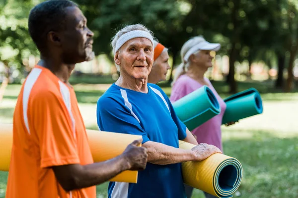 Selective focus of happy multicultural senior pensioners standing with fitness mats in park — Stock Photo