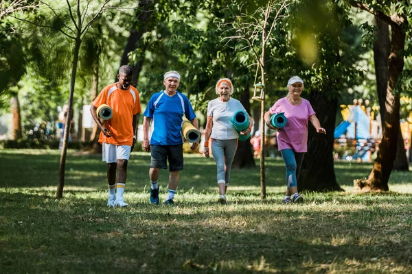 Enfoque selectivo de alegres pensionistas multiculturales y senior caminando con colchonetas de fitness en el parque - foto de stock