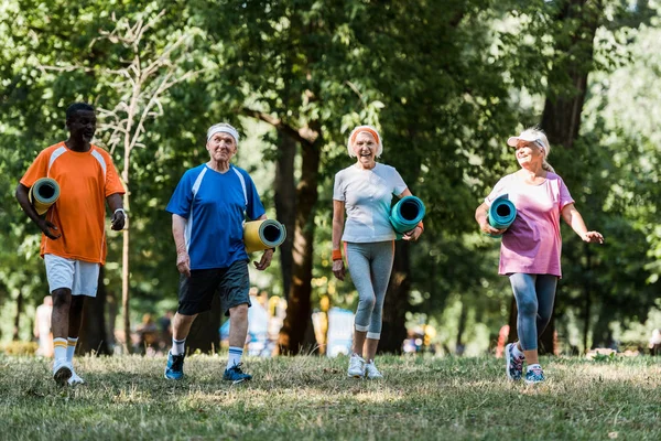 Positive and retired multicultural pensioners holding fitness mats and walking in park — Stock Photo