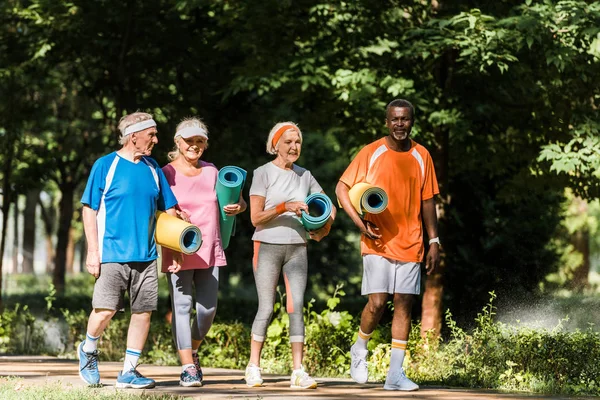 Positive retired and multicultural pensioners holding fitness mats and walking in park — Stock Photo