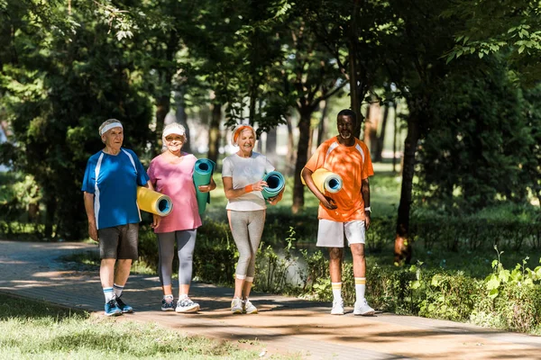 Cheerful retired and multicultural pensioners holding fitness mats and walking in park — Stock Photo