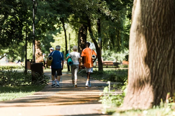 Selective focus of senior and multicultural pensioners holding fitness mats and walking in park — Stock Photo