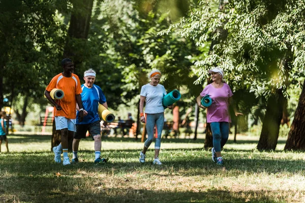 Избирательный фокус счастливых пожилых и мультикультурных пенсионеров, держащих спортивные коврики и ходящих по траве — стоковое фото