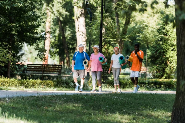 Foyer sélectif des retraités âgés et multiculturels gais tenant des tapis de fitness et marchant dans le parc — Photo de stock