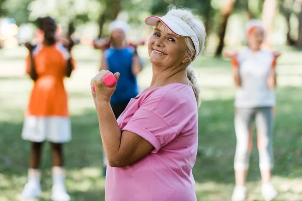 Selektiver Fokus einer fröhlichen Rentnerin mit Mütze, die beim Sport in der Nähe von Rentnern Hanteln hält — Stockfoto