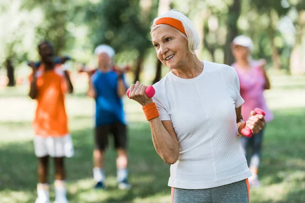 Selektiver Fokus der fröhlichen Seniorin, die beim Sport in der Nähe von Rentnern Hanteln hält — Stockfoto