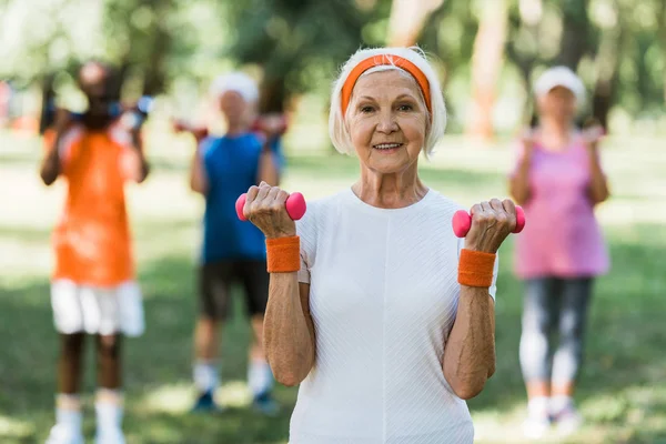 Mujer mayor sosteniendo pesas mientras hace ejercicio cerca de los pensionistas en el parque - foto de stock