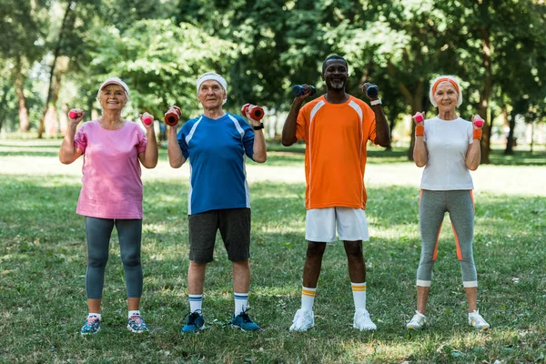 Alegres pensionistas multiculturales en ropa deportiva haciendo ejercicio con pesas en el parque - foto de stock