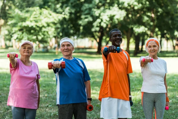 Jubilados multiculturales sonrientes haciendo ejercicio con mancuernas en el parque - foto de stock