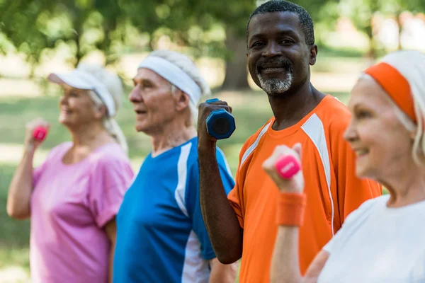 Enfoque selectivo del hombre afroamericano feliz ejercitarse con pesas con los pensionistas en el parque - foto de stock