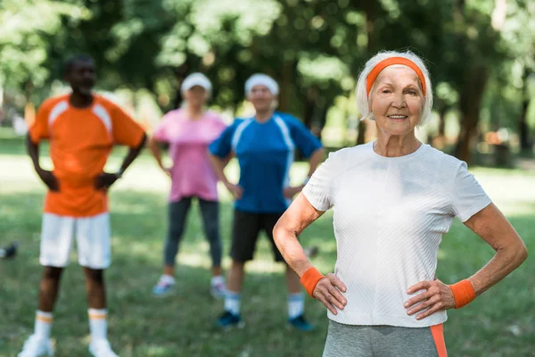 Selective focus of cheerful senior woman standing with hands on hips near multicultural pensioners in park — Stock Photo