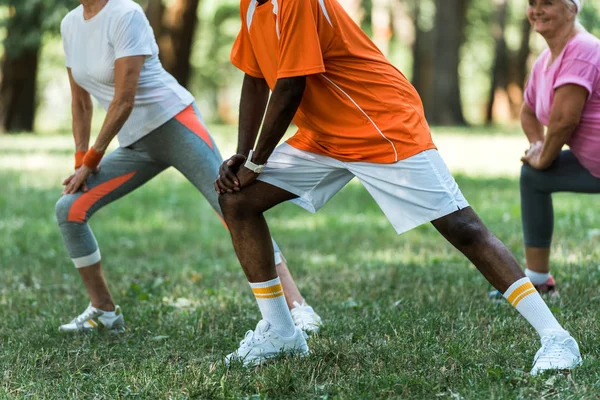 Cropped view of milticultural retired women and man stretching on grass in park — Stock Photo
