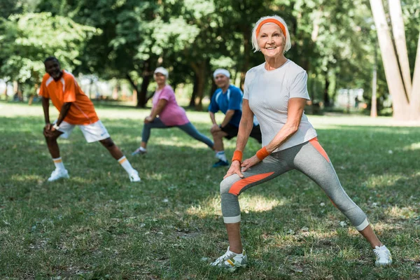 Selective focus of cheerful retired woman stretching near multicultural pensioners on grass — Stock Photo