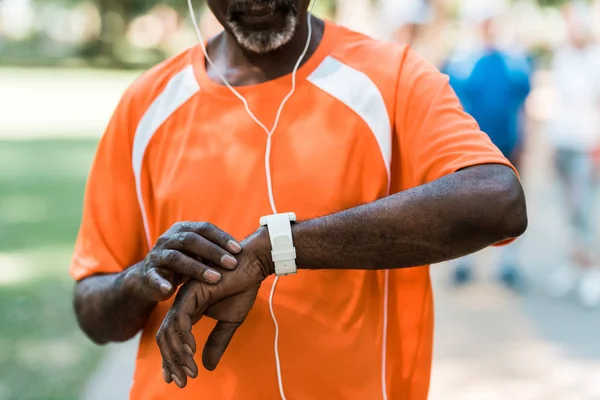 Vista ritagliata di anziano uomo africano americano in auricolari toccare orologio intelligente — Foto stock