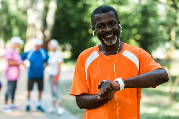 Selective focus of happy senior african american man in earphones touching smart watch — Stock Photo