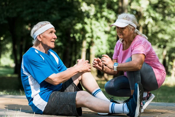 Pensionato uomo toccare ginocchio mentre seduto sul marciapiede vicino anziano donna in cap — Foto stock