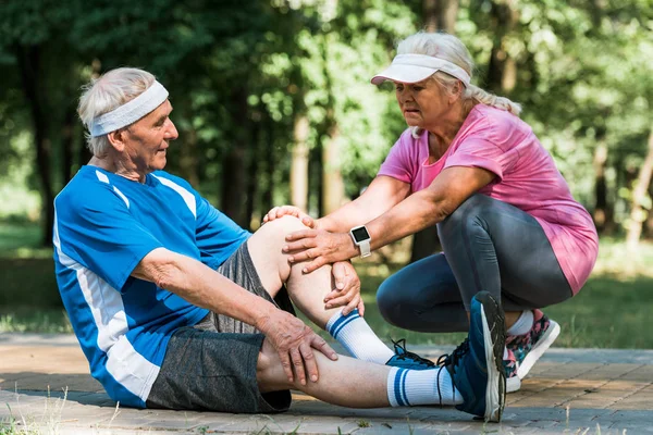 Retired upset woman touching knee of senopr man while sitting on walkway in park — Stock Photo