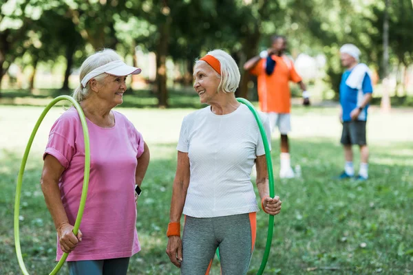 Enfoque selectivo de mujeres mayores felices sonriendo mientras sostiene aros de hula cerca de hombres jubilados multiculturales - foto de stock