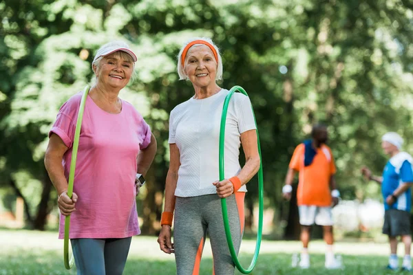 Selective focus of sportive senior women smiling while holding hula hoops near multicultural retired men — Stock Photo