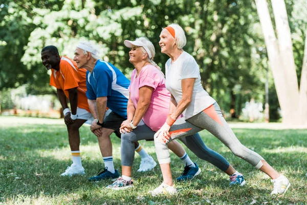 Foco seletivo de pessoas aposentadas e multiculturais felizes fazendo exercício de alongamento na grama — Fotografia de Stock