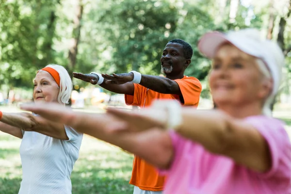 Enfoque selectivo del hombre afroamericano haciendo ejercicio cerca de las mujeres jubiladas en el parque - foto de stock