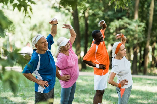 Enfoque selectivo de hombres y mujeres jubilados multiculturales alegres de pie con las manos en las caderas mientras hace ejercicio — Stock Photo