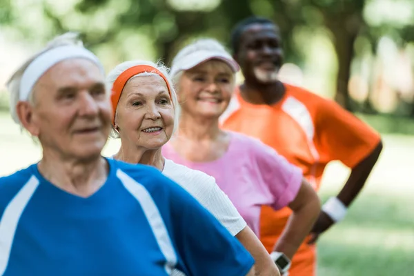 Selective focus of cheerful retired woman standing with hands on hips with multicultural pensioners — Stock Photo