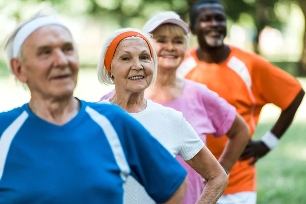 Избирательный фокус веселой пенсионерки, стоящей с руками на бедрах с мультикультурными пенсионерами в спортивной одежде — стоковое фото