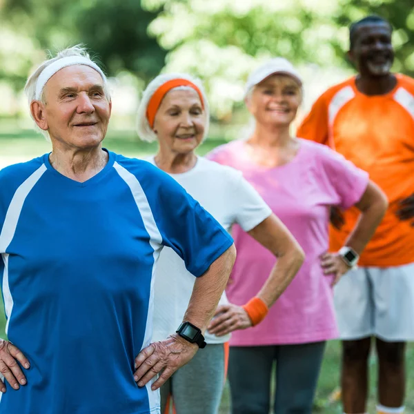 Selective focus of cheerful retired man standing with hands on hips with multicultural pensioners in sportswear — Stock Photo