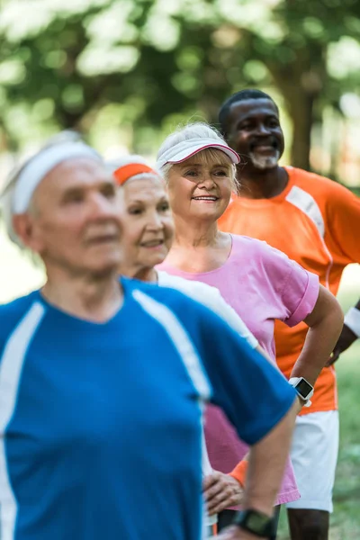 Messa a fuoco selettiva di allegro pensionato afroamericano in piedi con le mani sui fianchi con i pensionati in abbigliamento sportivo — Foto stock