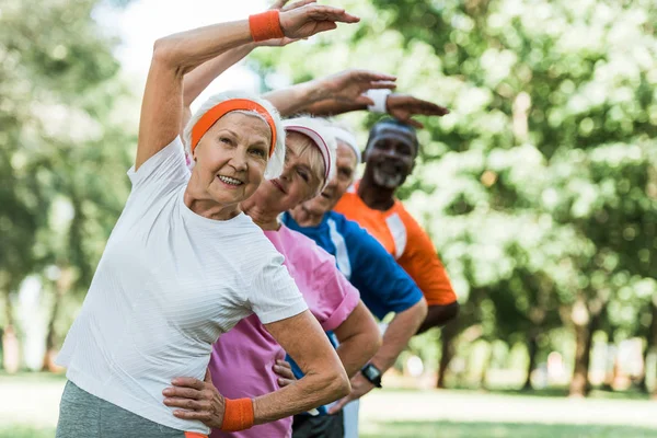 Селективное внимание веселой пенсионерки рядом с мультикультурными мужчинами и женщинами, делающими упражнения на растяжку — стоковое фото