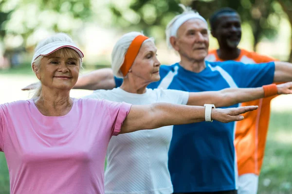 Selective focus of happy retired woman standing with outstretched hands near multicultural pensioners — Stock Photo