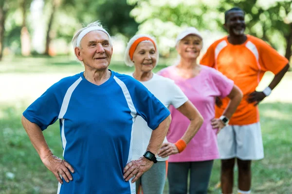 Selective focus of retired man standing with hands on hips with multicultural pensioners — Stock Photo