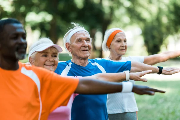 Selective focus of happy retired man standing with outstretched hands near multicultural pensioners in park — Stock Photo