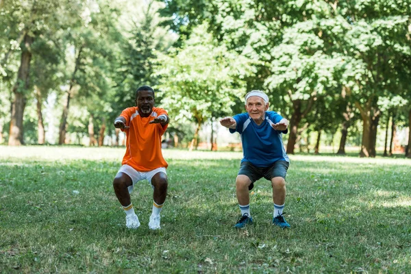Щасливі старші та мультикультурні медики сидять на траві біля дерев — стокове фото