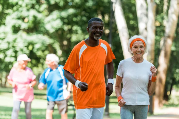 Selective focus of african american senior man and happy retired woman exercising near pensioners outside — Stock Photo