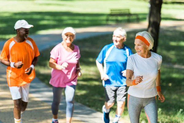 Foco seletivo de mulher aposentada feliz correndo com aposentados multiculturais no parque — Fotografia de Stock