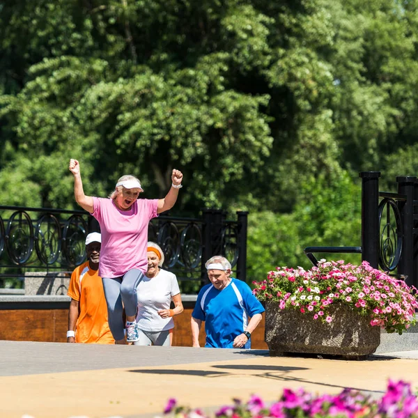 Foco seletivo de mulher aposentada alegre celebrando triunfo perto de pensionistas multiculturais no parque — Fotografia de Stock
