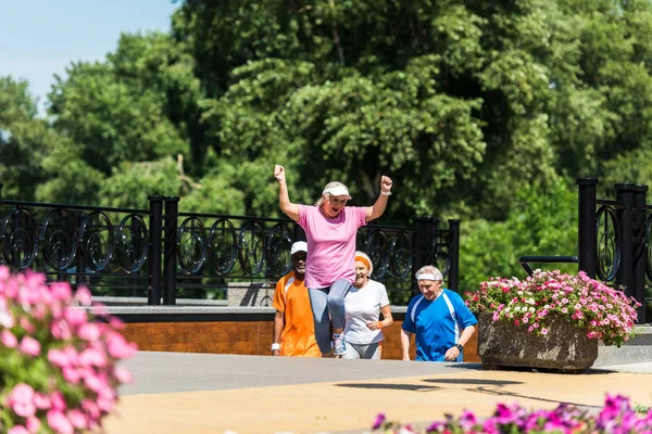 Selective focus of happy retired woman celebrating triumph near multicultural pensioners in park — Stock Photo