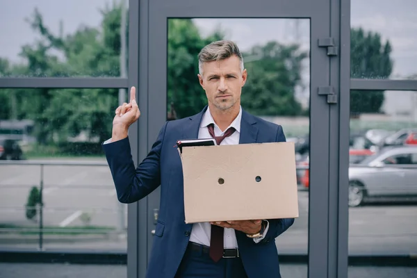 Hombre guapo en traje mostrando el dedo medio mientras sostiene la caja cerca del edificio - foto de stock